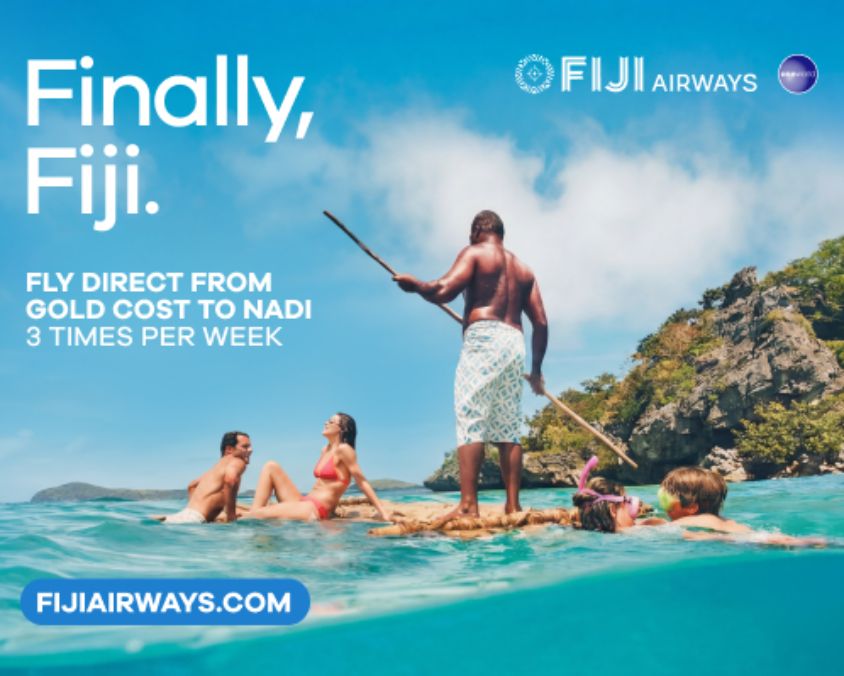Promotional image for Fiji Airways showing a group of travellers enjoying a tropical ocean setting in Fiji. A local guide stands on a traditional raft in clear turquoise water, with lush green cliffs and blue skies in the background. Text reads: “Finally, Fiji. Fly direct from Gold Coast to Nadi, 3 times per week.” Fiji Airways branding and website URL are displayed.