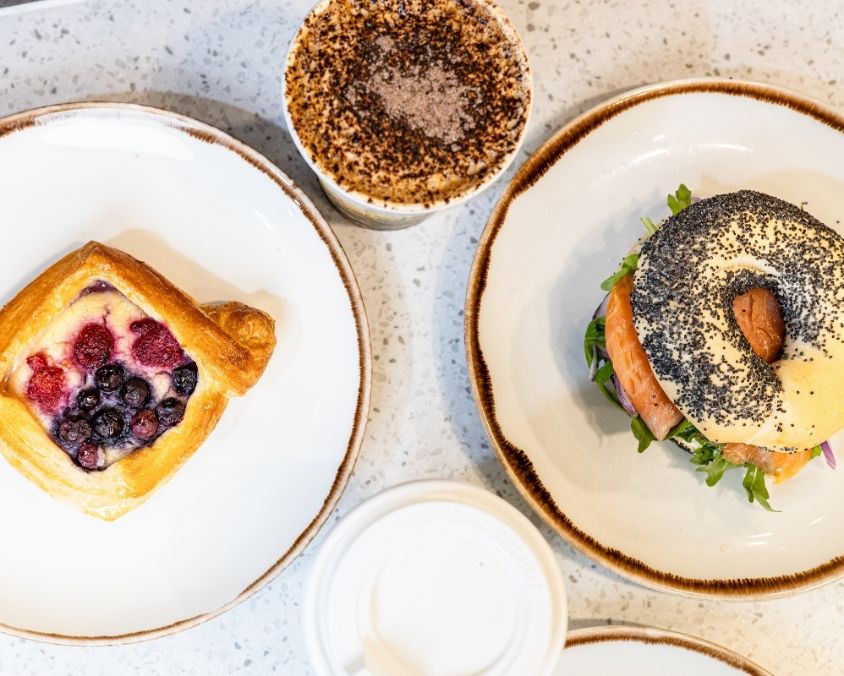 A top-down view of a café-style meal featuring a berry danish pastry, a seeded bagel sandwich with leafy greens, a takeaway coffee, and a white coffee cup lid on a speckled tabletop.