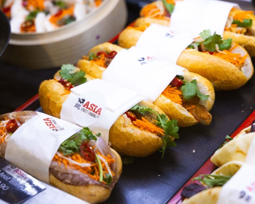 A display of freshly made Vietnamese bánh mì rolls, filled with ingredients like sliced meats, pickled vegetables, coriander, and chilli, wrapped in branded “Taste Asia” paper on a serving tray.