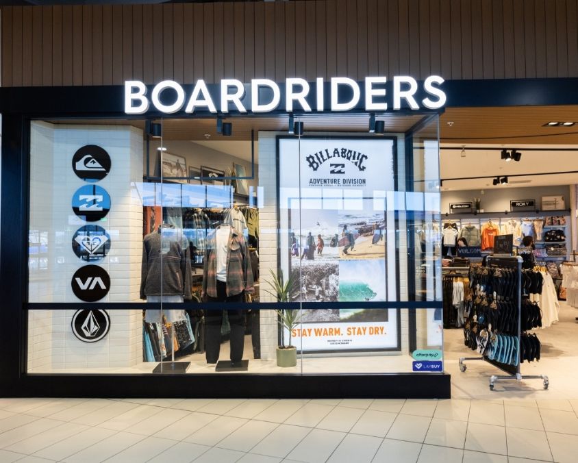 The exterior of the Boardriders retail store at an airport, featuring a bright storefront with surf and skate brand logos on the window and displays of casual clothing and gear inside.