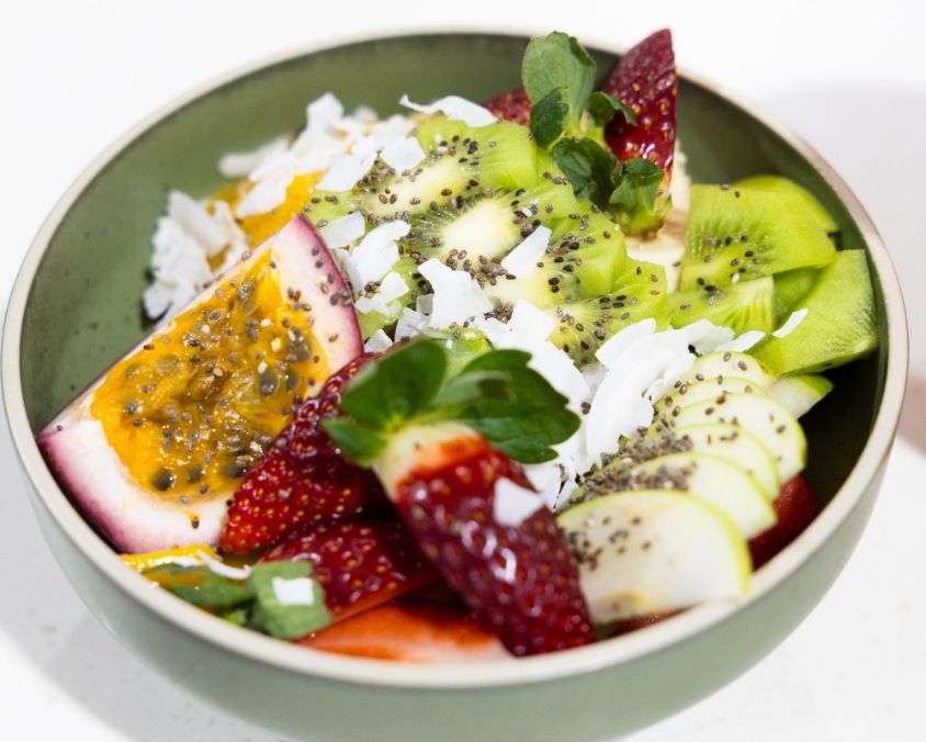 A vibrant fruit bowl filled with sliced strawberries, kiwi, banana, passionfruit, shredded coconut, chia seeds, and fresh greens, served in a green ceramic bowl.