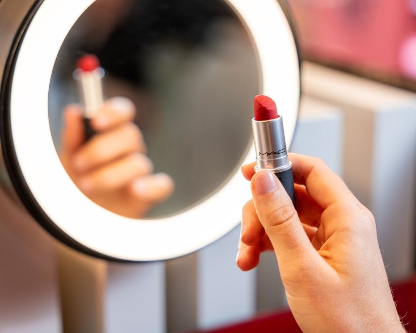A close-up of a person holding a red lipstick in front of a round, illuminated mirror, with the reflection of the lipstick visible in the mirror.