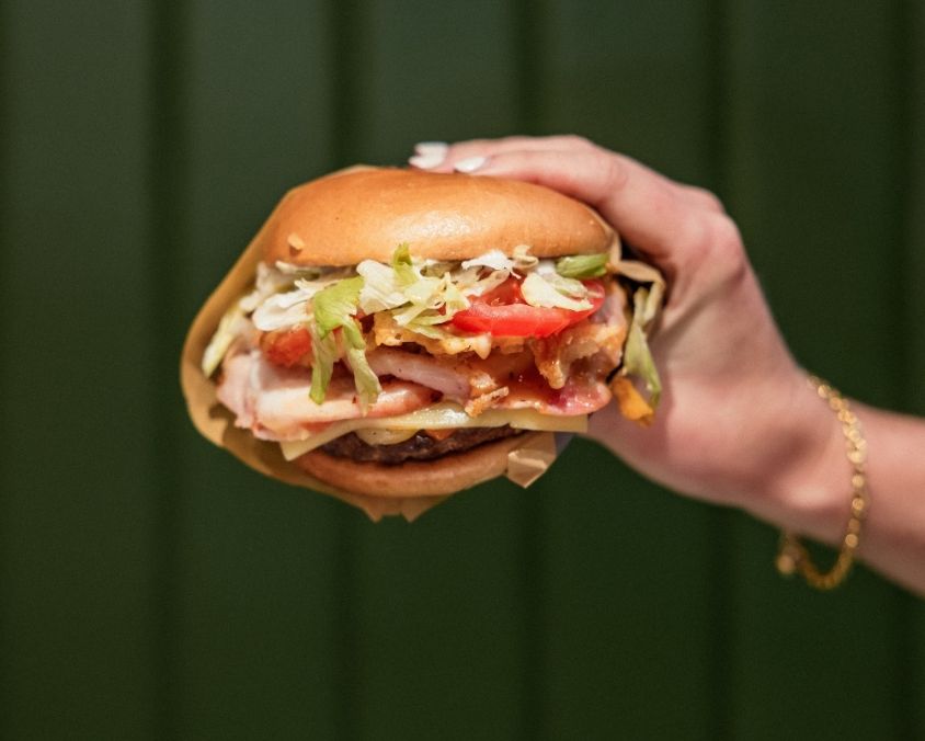  A hand holding a large, juicy burger filled with lettuce, tomato, pickles, bacon, cheese, and sauce, in a glossy toasted bun, against a dark green background.