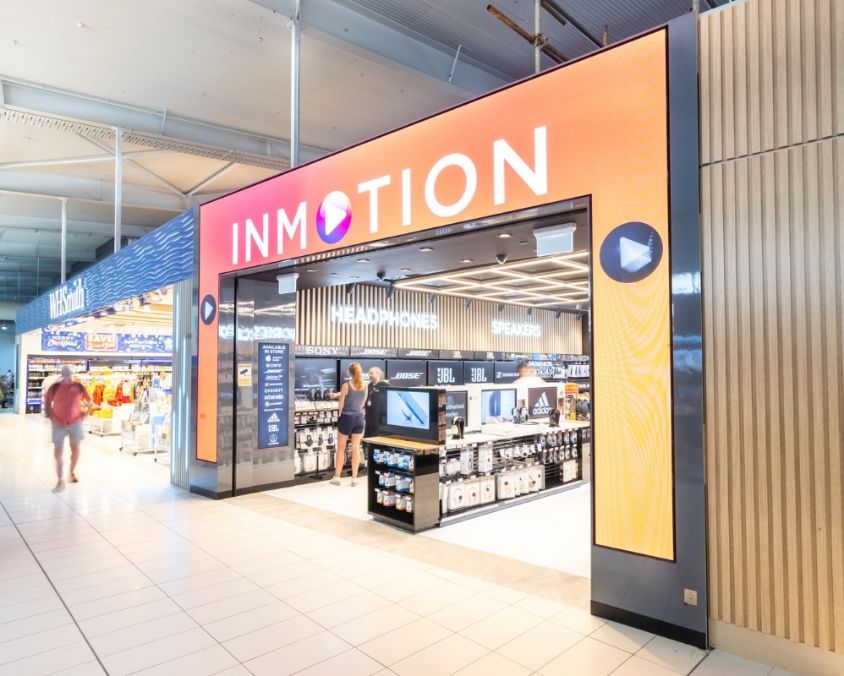 The exterior of the InMotion store at an airport, featuring a bold orange and pink storefront with tech accessories and electronics on display inside, and passengers walking nearby.