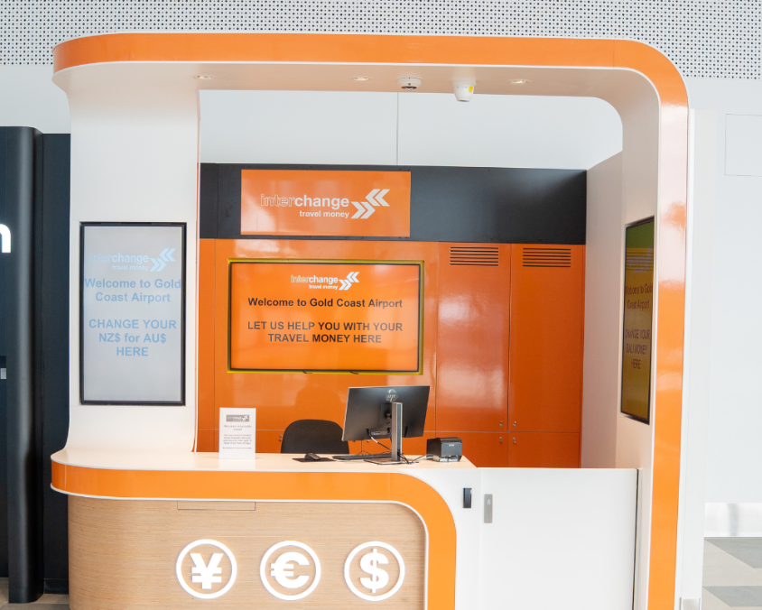 Interchange Travel Money currency exchange booth at Gold Coast Airport, with bright orange and white signage displaying messages about exchanging NZ$ for AU$, a counter with a computer, and currency symbols for yen, euro, and dollar.