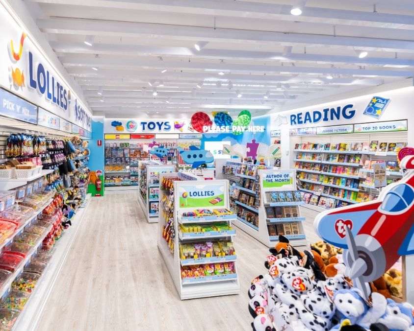 A brightly lit children's store at the airport featuring colourful signage for lollies, toys, and reading. Shelves are stocked with books, plush toys, games, and confectionery, creating a playful and inviting space for young travellers.