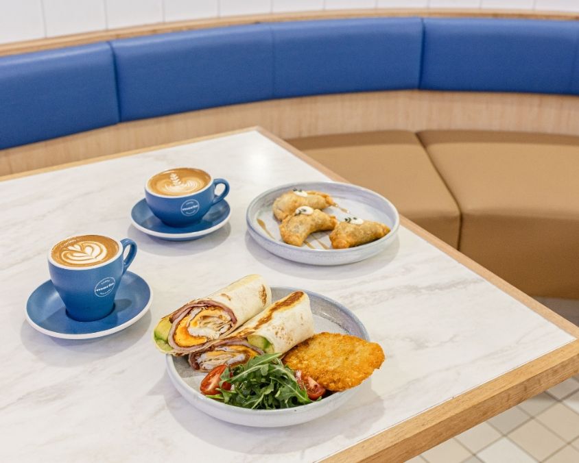 A cosy café setting with a marble table featuring two blue cups of coffee, a breakfast wrap with hash browns and salad, and a plate of pastries. Blue and tan booth seating is visible in the background.