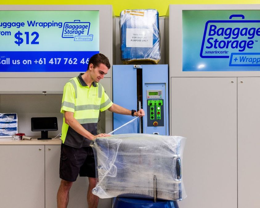 A staff member in a high-visibility shirt wraps luggage in plastic at a baggage storage and wrapping station. Signs above display pricing and contact details for the service.
