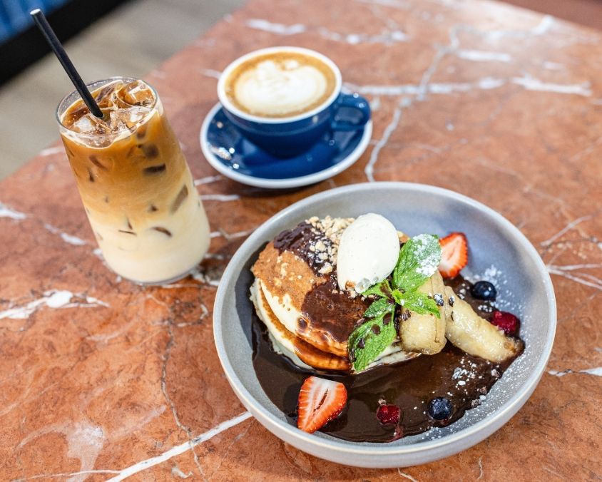 A marble café table set with a plate of pancakes topped with whipped cream, banana, berries, and chocolate sauce, alongside an iced latte and a hot coffee in a blue cup.