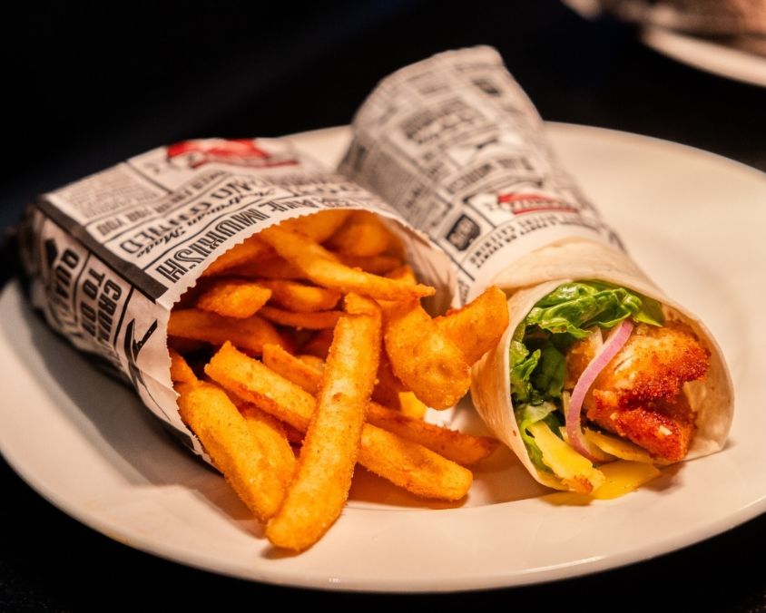 A white plate with a wrap and golden chips from Schnitz. The wrap is filled with crispy chicken schnitzel, lettuce, red onion, and sauce, all wrapped in newspaper-style paper.