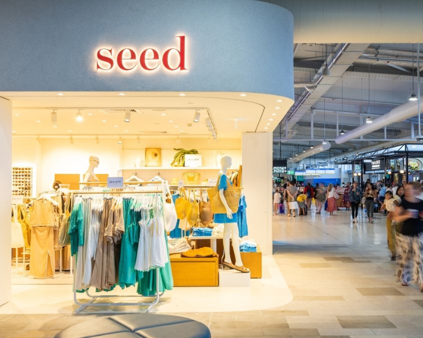 The exterior of the Seed Heritage store at an airport, showcasing a bright and modern storefront with mannequins dressed in stylish neutral and turquoise outfits, and travellers walking past in the terminal.
