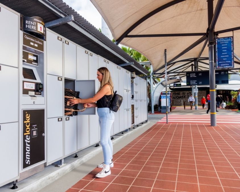 A woman uses a Smarte Carte luggage locker system at an outdoor terminal area. She is placing or retrieving a bag from a secure compartment under a shaded walkway with signs and people in the background.
