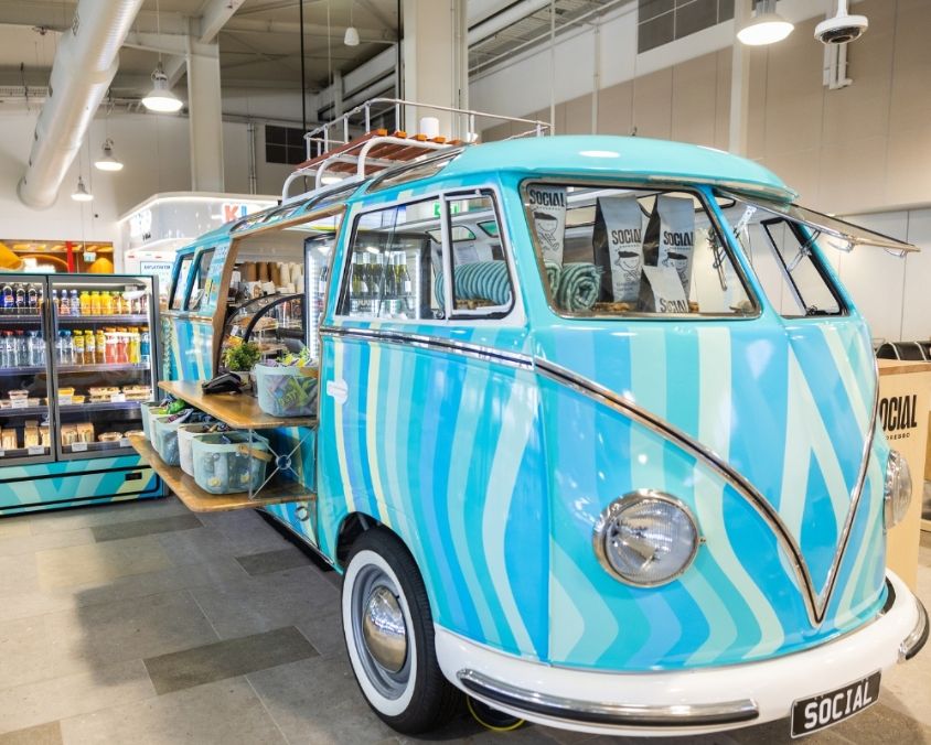 A retro turquoise and blue striped van repurposed as a retail display inside a store, featuring surf-style homewares and beach accessories arranged on open shelves extending from the van.