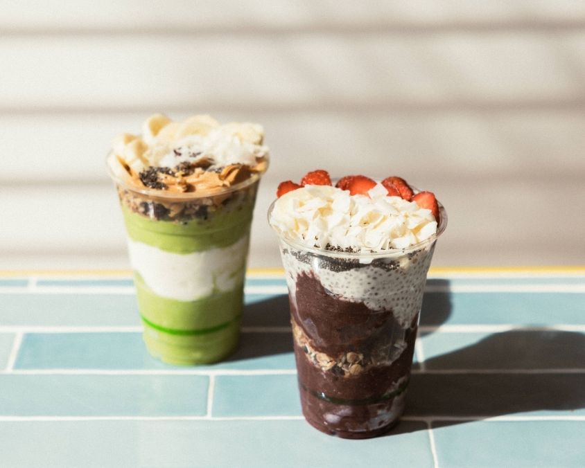 Two layered smoothie cups placed on a tiled surface in sunlight. One features green and white layers topped with banana slices and granola, while the other has dark purple açaí, strawberries, coconut flakes, and granola.