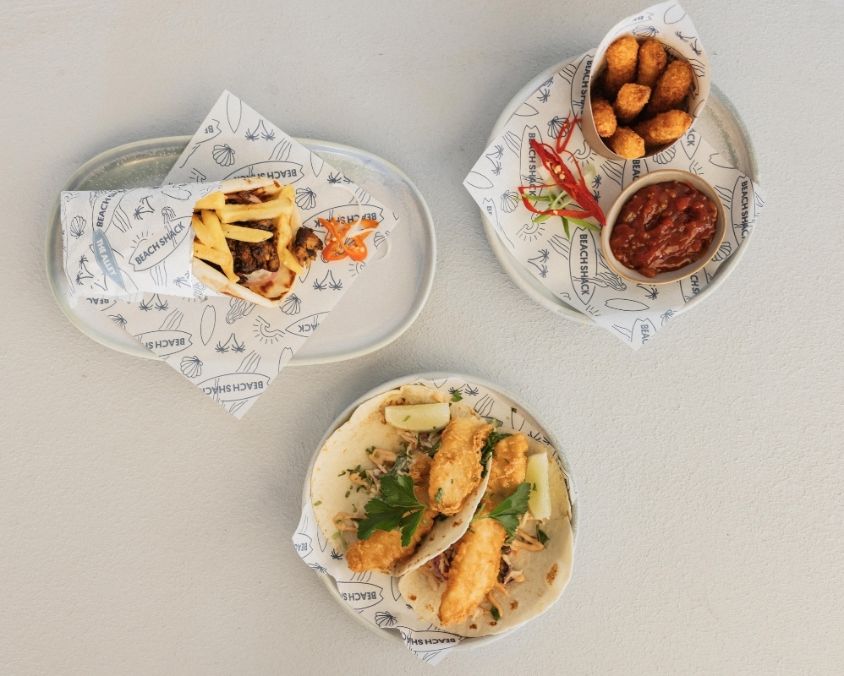  A flat lay of three Mediterranean-inspired dishes, including a pita wrap with grilled meat and fries, crispy golden bites served with red dipping sauce and chilli, and a plate of halloumi garnished with herbs.