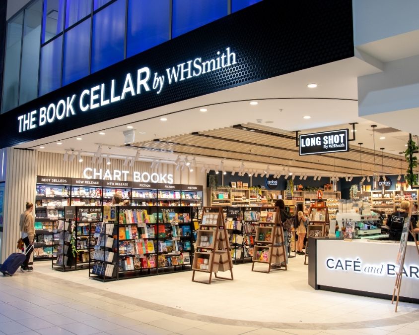 The exterior of The Book Cellar by WHSmith at an airport, featuring neatly arranged book displays, a café counter, and signage for chart books and grab-and-go coffee. Travellers browse the space in a bright, welcoming setting.