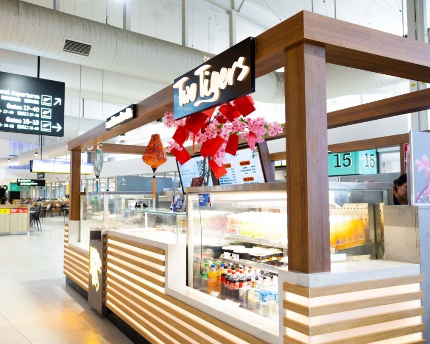 The Two Tigers food kiosk at an airport, featuring a modern wood-accented design with illuminated panels, red lantern decorations, and a fridge stocked with bottled drinks. A departure gate display is visible in the background.