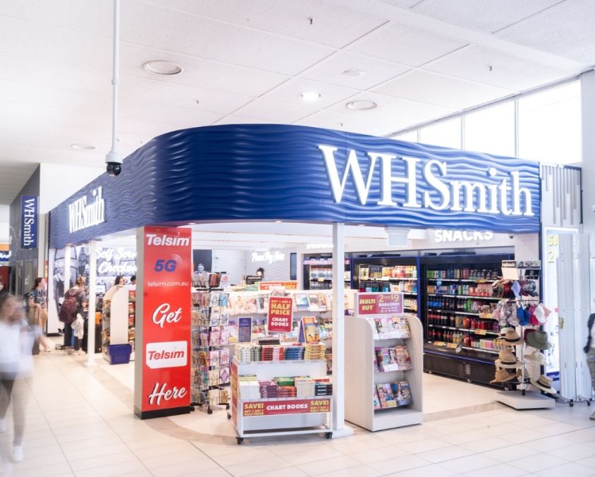 The exterior of a WHSmith store at an airport, featuring bright signage, promotional displays, and shelves stocked with books, snacks, drinks, and travel essentials. Passengers walk by in the terminal corridor.