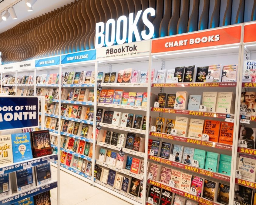  A bright and well-organised book section inside WHSmith at the airport, featuring shelves labelled with categories like BookTok, New Fiction, and Chart Books. A display stand highlights the Book of the Month.