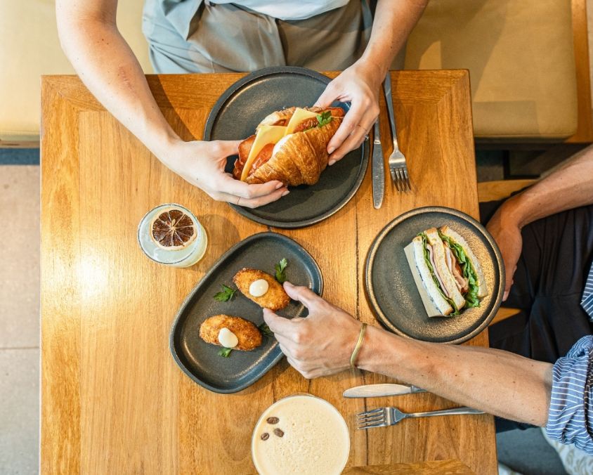 A top-down view of two people dining at a wooden café table, enjoying a croissant sandwich, a club sandwich, arancini balls, and iced coffees, served on dark ceramic plates.