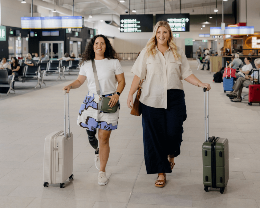 Two women walk confidently through Gold Coast Airport with carry-on luggage. One woman has a prosthetic leg, and both are smiling as they move through the bright, modern terminal.