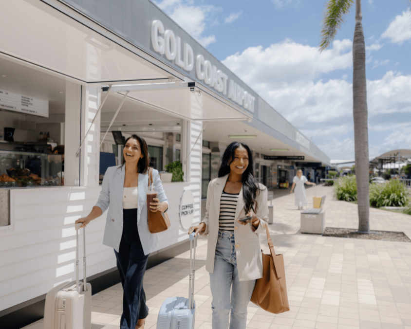 Two women walk outside Gold Coast Airport, smiling with carry-on luggage and handbags. A café and palm trees frame the sunny, relaxed atmosphere of the terminal exterior.