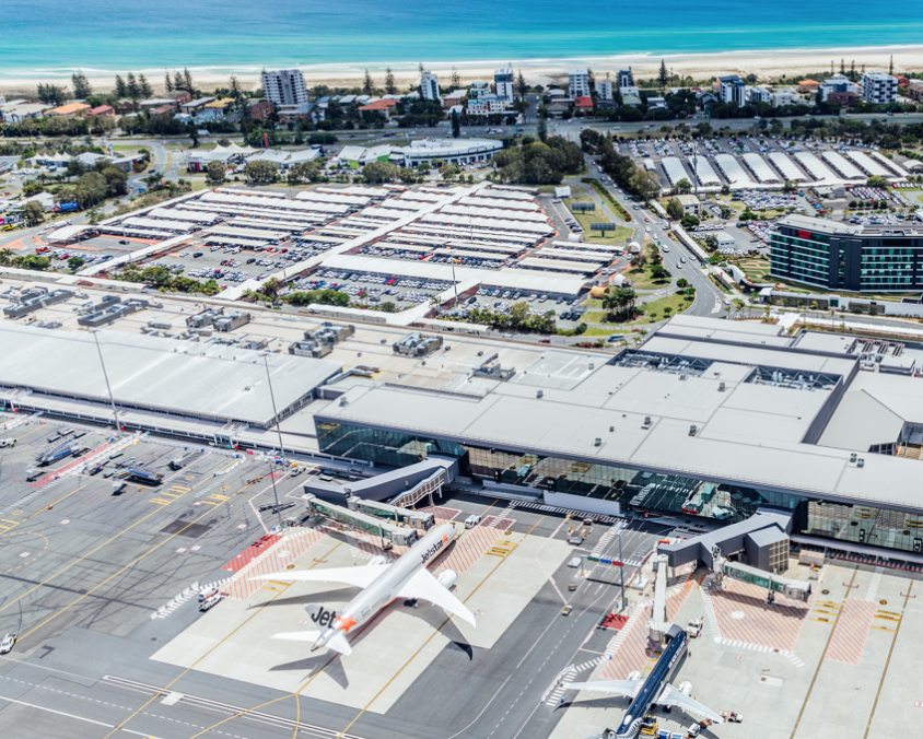 Aerial view of Gold Coast Airport with aircraft parked at the terminal gates, car parks and terminal infrastructure in the midground, and the beach and ocean stretching out in the distance.