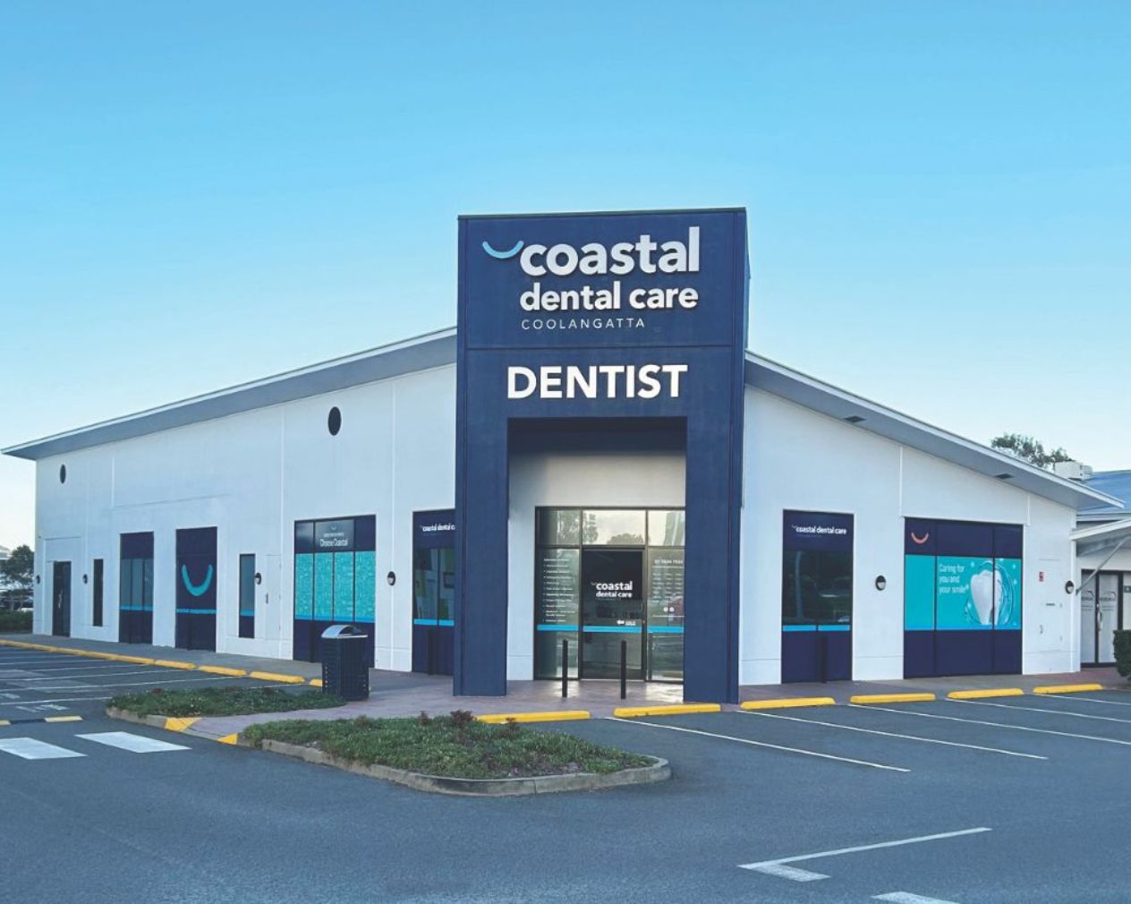 Exterior view of Coastal Dental Care Coolangatta, a modern white building with blue signage reading 'Dentist' above the entrance, surrounded by parking spaces and a clear blue sky.