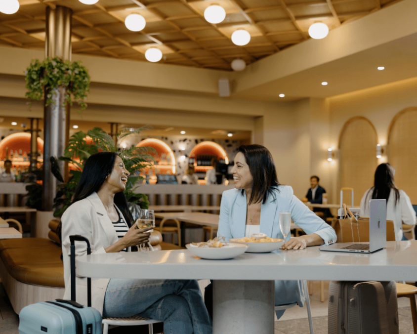 Two travelers seated at a modern dining area inside Gold Coast Airport, enjoying drinks and snacks with luggage beside the table.