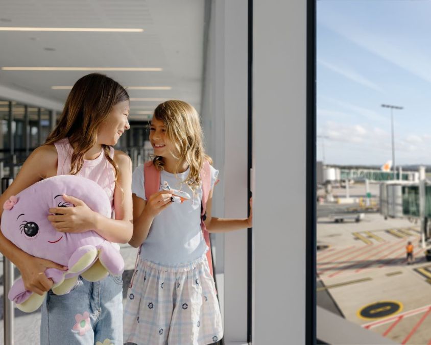 Two children standing inside an airport terminal near large windows overlooking the tarmac, holding a plush toy and wearing backpacks.
