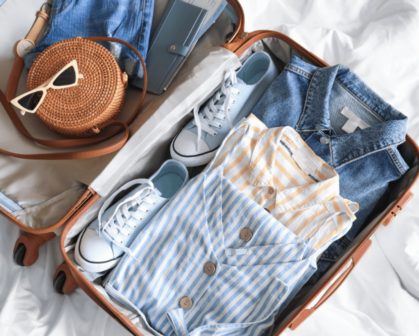 Open suitcase neatly packed with striped shirts, denim jacket, white sneakers, round wicker bag, sunglasses, and travel wallet on a bed.