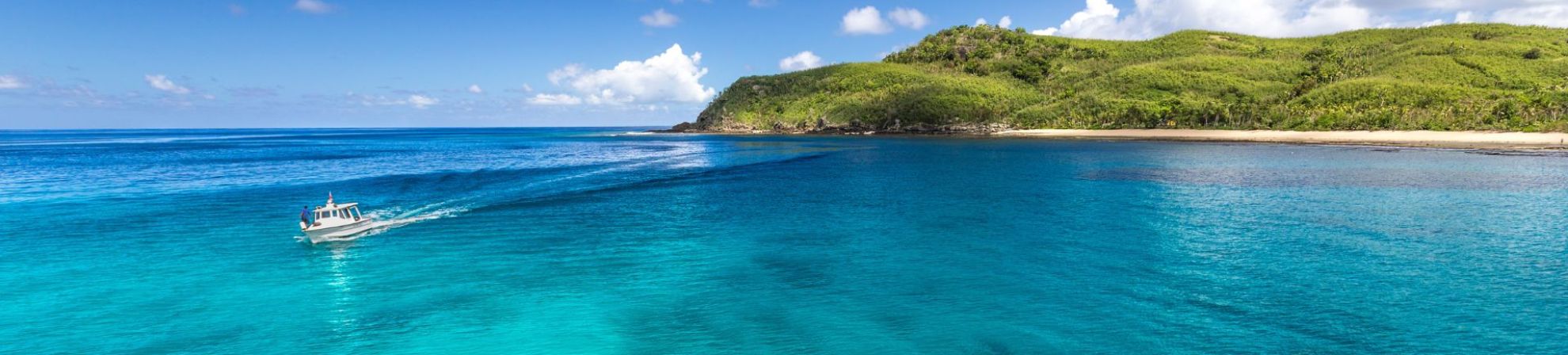 A small boat travels across bright turquoise water near a tropical island coastline with lush green hills under a blue sky.