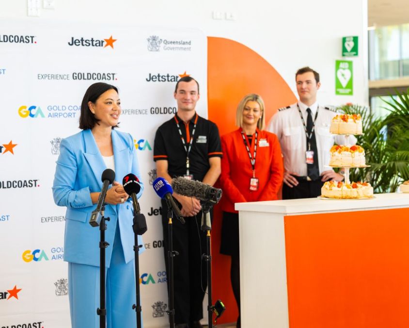 Press event at Gold Coast Airport with Jetstar branding, featuring speakers and staff standing near microphones and a dessert display