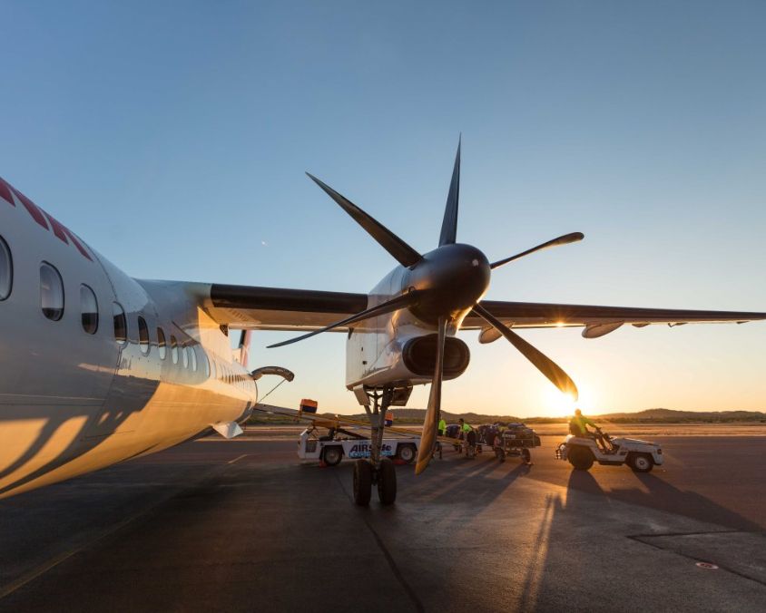 Propeller aircraft on airport tarmac during sunset, with ground support equipment visible in the background.