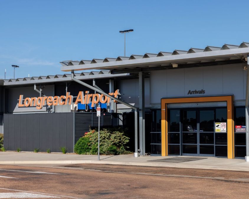 Exterior view of Longreach Airport entrance with signage for "Longreach Airport" and "Arrivals"; modern building with metal roof and greenery under a clear blue sky.