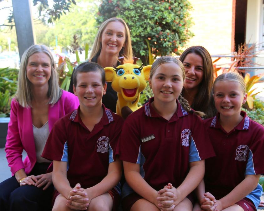 Three adults and three children pose in front of a garden area with various plants. The children wear matching maroon shirts with blue accents and an emblem, while one adult holds a yellow giraffe plush toy. Behind them is a building with windows displaying papers.