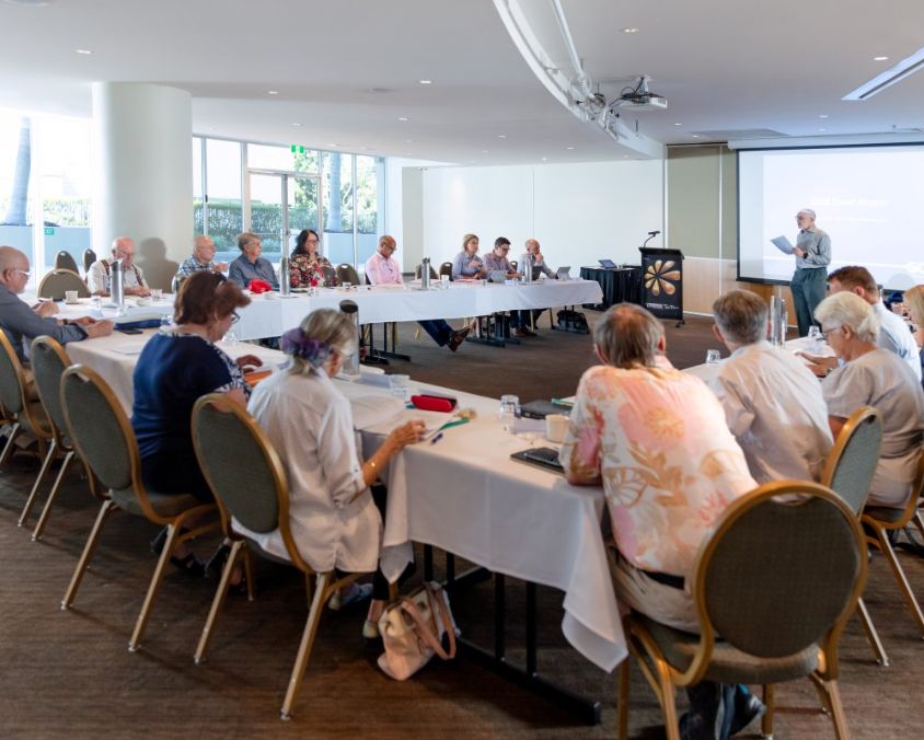 Community consultation meeting held in a conference room with around 20 attendees seated in a U-shaped table arrangement. One person stands at the front near a projector screen, leading the discussion in a formal and professional setting