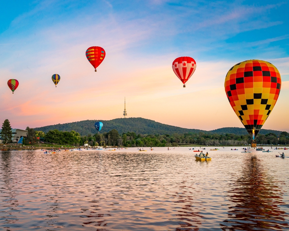 Colourful hot air balloons float above a lake at sunrise in Canberra, Australia. Reflections dance on the water as kayakers paddle below, with Telstra Tower visible atop the hill in the background.