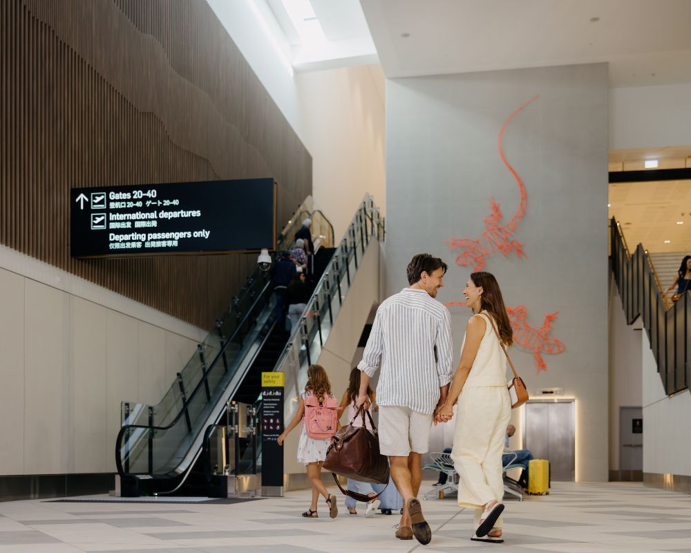 A family walks through Gold Coast Airport past the red gecko wall art and escalators, making their way towards international departures.