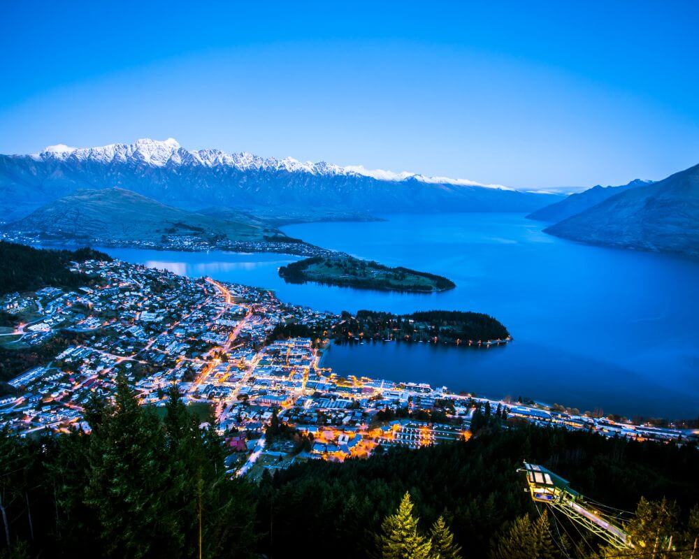 A twilight view of Queenstown, New Zealand, with the town’s lights glowing beside Lake Wakatipu. Snow-capped mountains of The Remarkables range rise in the distance under a deep blue sky.