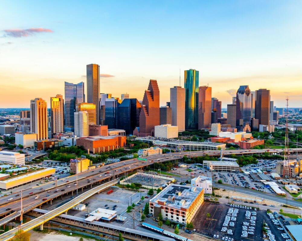 Aerial view of downtown Houston, Texas, at sunset. The skyline features a mix of modern skyscrapers and office towers, with elevated highways and busy roads weaving through the cityscape.