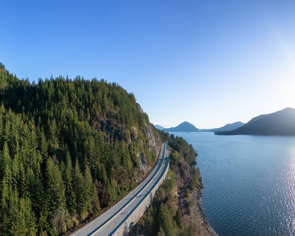 Aerial view of the Sea to Sky Highway near Vancouver, curving between lush green forest and the sparkling waters of Howe Sound, with mountains on the horizon.