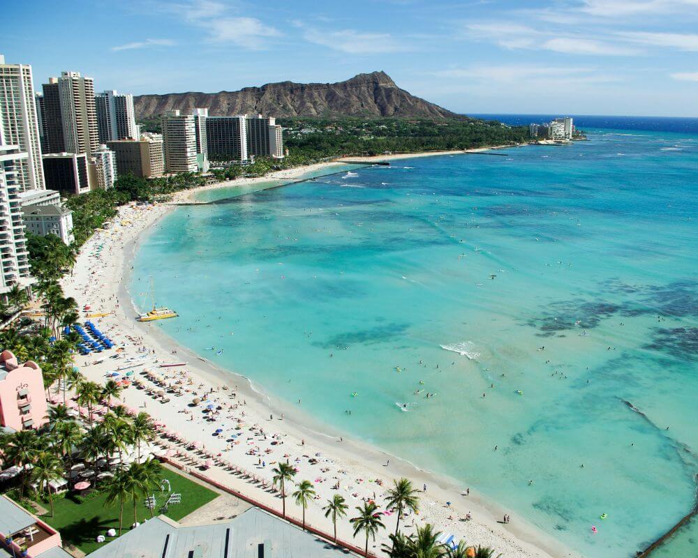Aerial view of Waikiki Beach in Honolulu, Hawaii, featuring turquoise waters, high-rise hotels, palm trees, and the iconic Diamond Head crater in the background. The beach is lined with sunbathers and swimmers enjoying the tropical coastline.