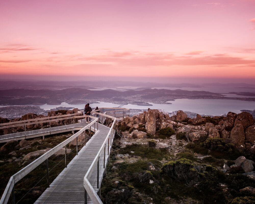 Elevated walkway on kunanyi / Mount Wellington overlooking Hobart, Tasmania, at sunset. The sky glows pink and purple as visitors take in panoramic views of the city, harbour, and surrounding landscape.