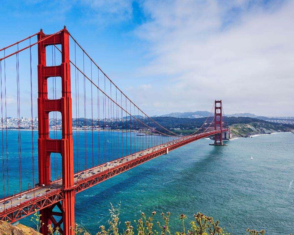 The Golden Gate Bridge in San Francisco, California, stretches across the bay under a partly cloudy sky. The iconic red suspension bridge is surrounded by blue-green waters and rolling hills.