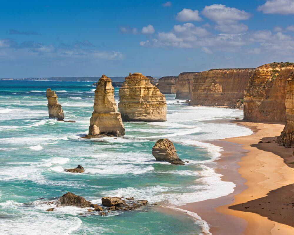 The Twelve Apostles along the Great Ocean Road in Victoria, Australia. Tall limestone stacks rise from the turquoise waters, with waves crashing against the cliffs and a sandy beach stretching along the coastline.