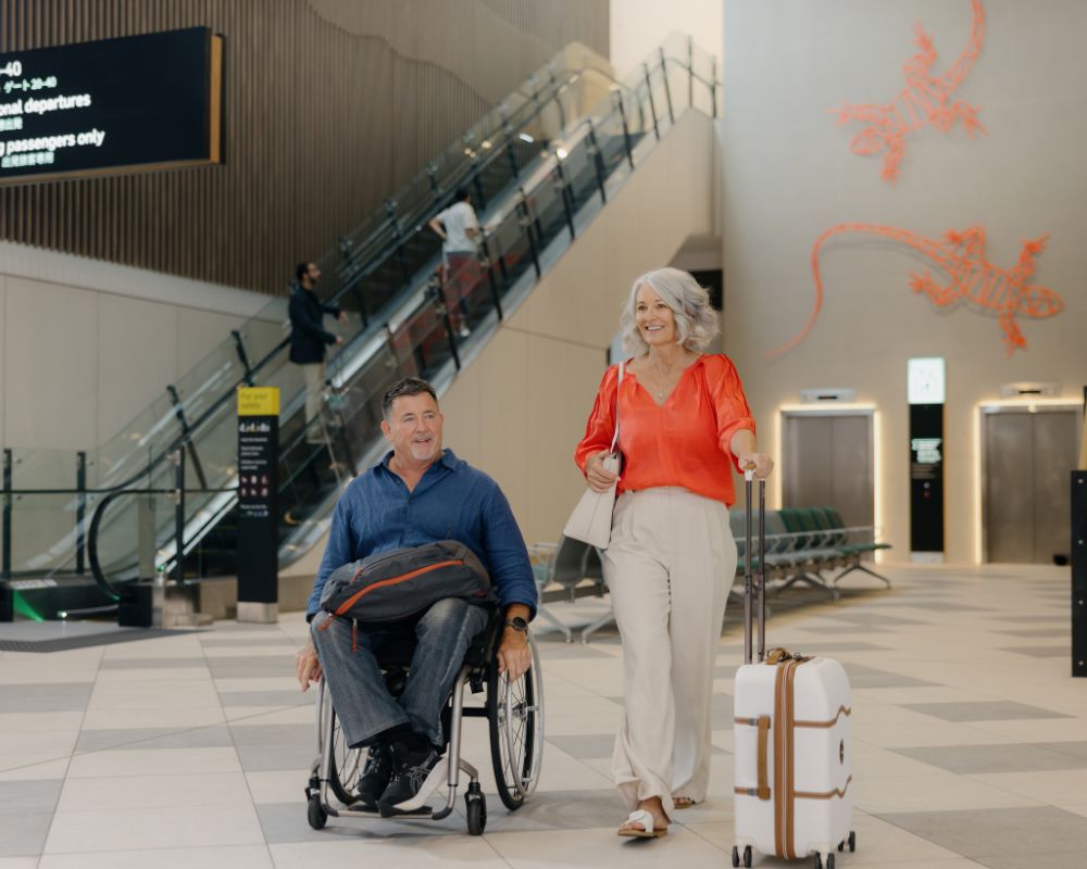  A man in a wheelchair and a woman with a suitcase walk together through a spacious airport terminal. Behind them are escalators, lifts, seating, and a red gecko wall mural, contributing to a bright, modern atmosphere.