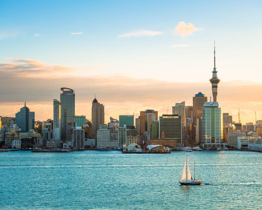 A scenic view of Auckland’s skyline at sunset, featuring the Sky Tower and waterfront buildings. A sailboat glides across the calm harbour waters in the foreground.