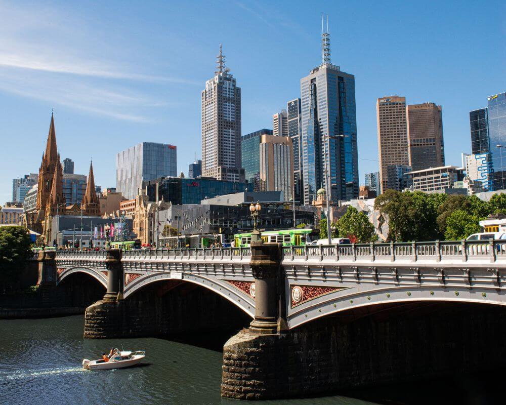 Scenic view of Melbourne’s city skyline with a mix of historic and modern buildings, including St Paul’s Cathedral. The Princes Bridge crosses the Yarra River, with a boat cruising beneath and a tram crossing above.
