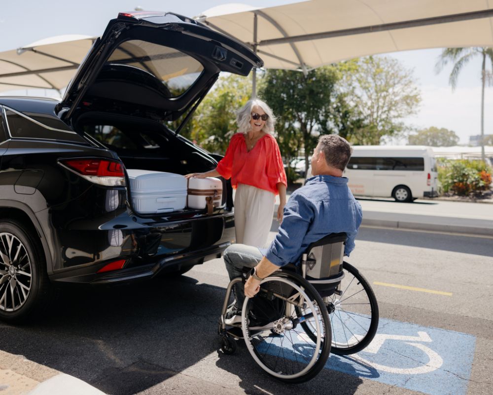 A man using a wheelchair and a woman retrieve luggage out of the boot of a car at Gold Coast Airport, with a shade sail visible behind them.